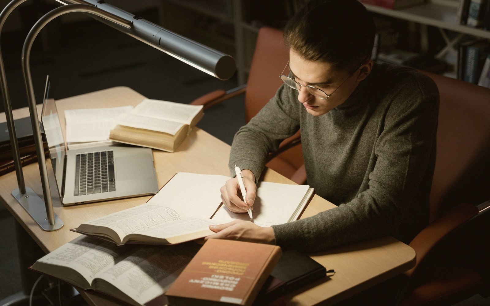 Man writing notes at a desk surrounded by open books and a laptop