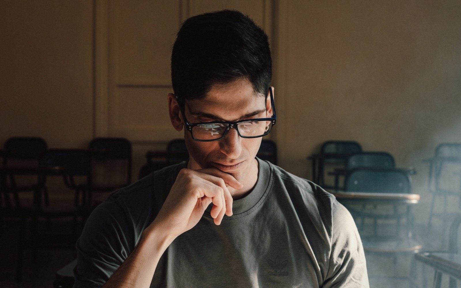 Young man in glasses rests chin on hand, looking down in an empty classroom